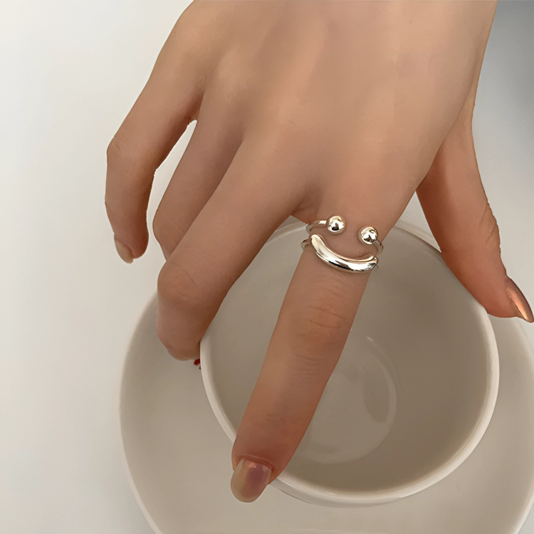 Main posée sur une tasse blanche, avec la bague sourire argentée à deux boules autour de l'index. 2