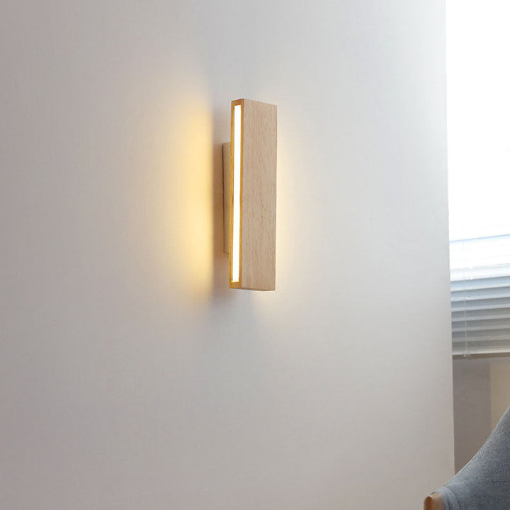 Applique en bois verticale allumée sur un mur blanc, dans une pièce avec store à lamelles et fauteuil gris. 15