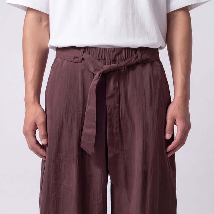Gros plan sur la taille élastique et la ceinture nouée du pantalon marron prune, tissu légèrement froissé. 9