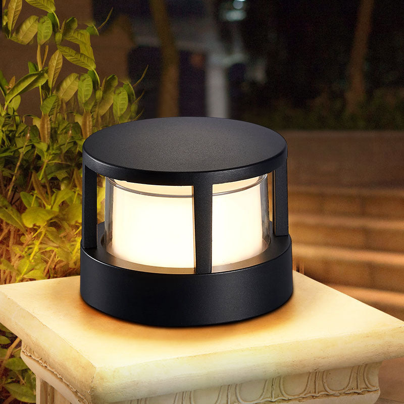 Lampe cylindrique noire à structure ajourée posée allumée sur un muret de pierre, devant un feuillage de jardin le soir. 1