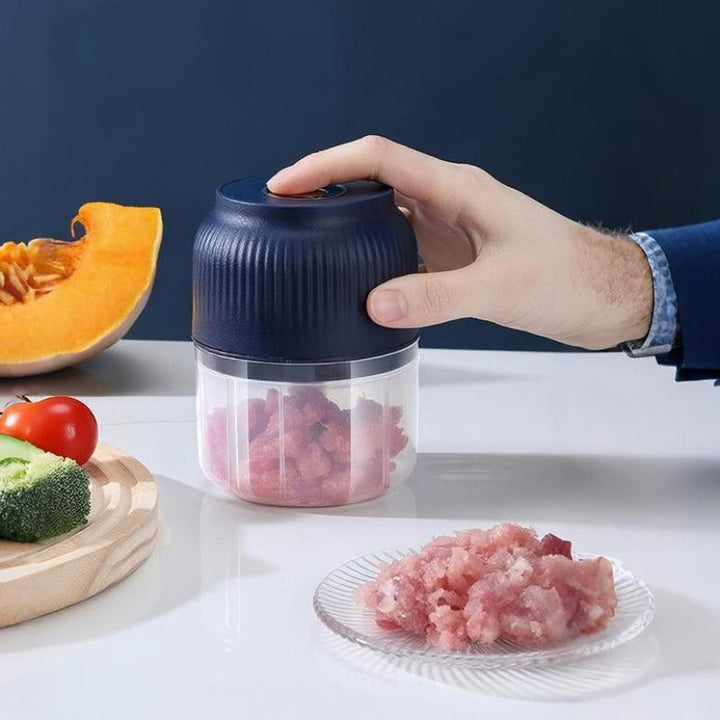 Une main appuie sur le mini hachoir bleu marine rempli de viande hachée, posé près d'une assiette de viande, d'un potiron et de brocoli. 1