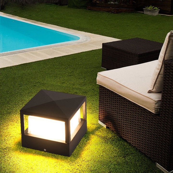 Lampe carrée à toit en pointe posée sur la pelouse, allumée, près d'une piscine et d'un fauteuil en rotin. 4