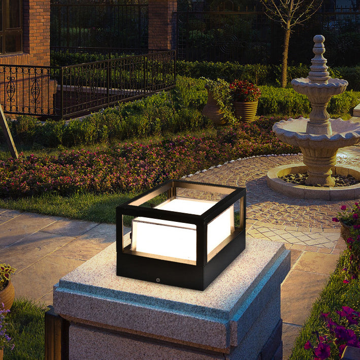 Lampe carrée noire à cadre ouvert, allumée, posée sur un pilier de pierre dans un jardin avec fontaine. 2