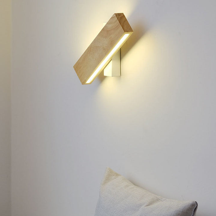 Applique en bois pivotée en diagonale sur un mur blanc, allumée, avec un coussin beige en bas de l'image. 1