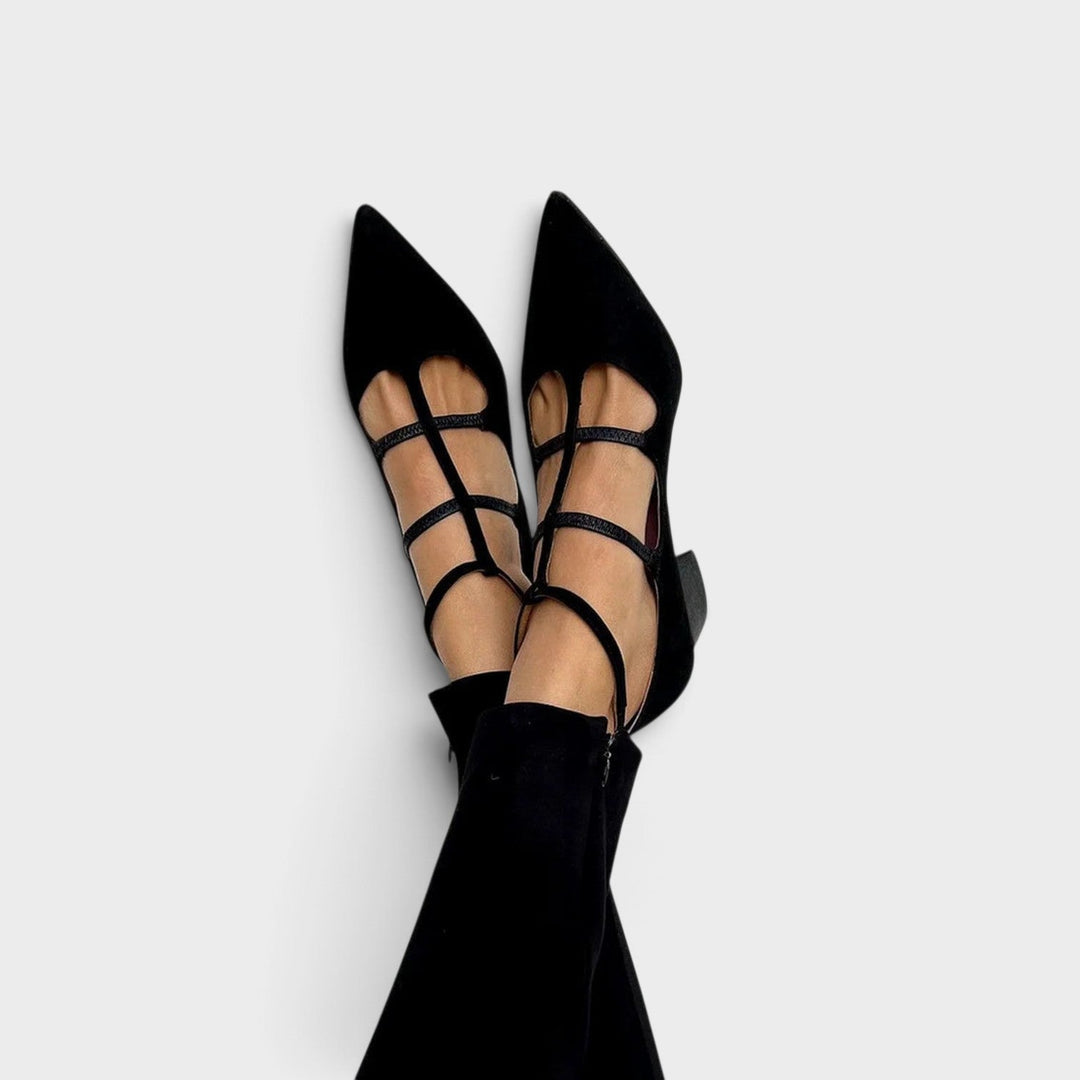 Jambes croisées en pantalon noir, pieds chaussés d'escarpins noirs à bout pointu, brides fines croisées sur le cou-de-pied et bride à la cheville. 1