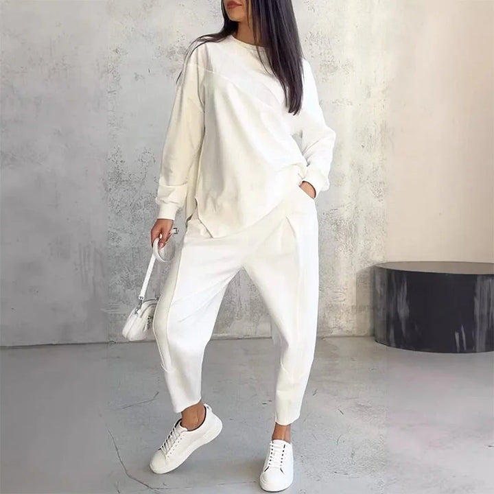 Femme en ensemble blanc, une main dans la poche, sac blanc à la main, baskets blanches, devant un mur en béton. 1