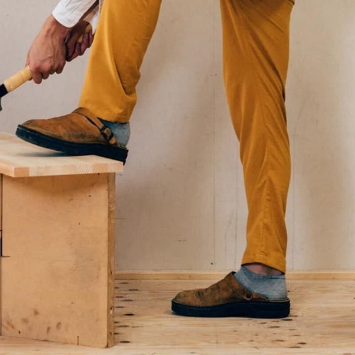 Personne en pantalon moutarde portant les mules brunes, un pied posé sur un banc en bois 3