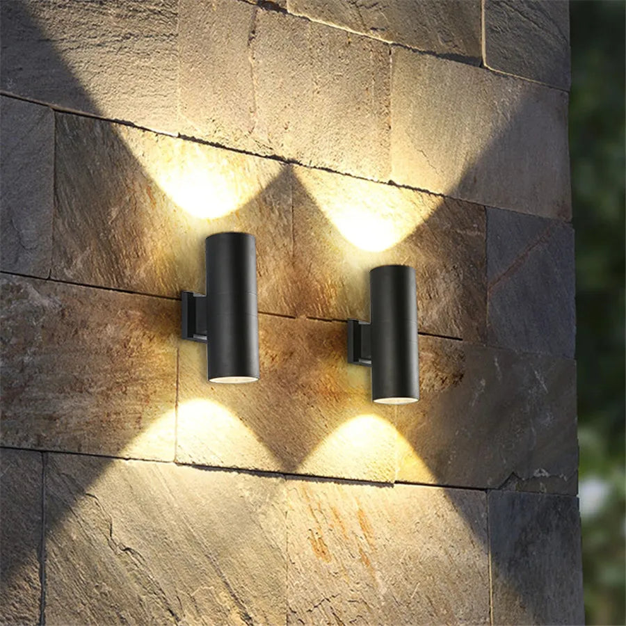 Deux appliques cylindriques noires allumées sur un mur de pierre, projetant des faisceaux de lumière chaude vers le haut et vers le bas. 2