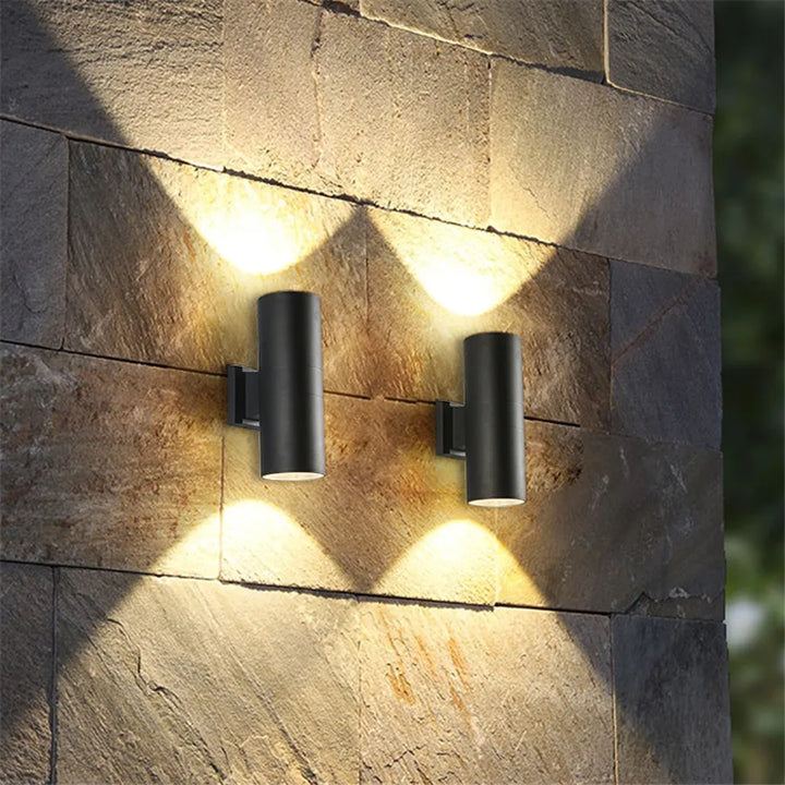 Deux appliques cylindriques noires allumées sur un mur de pierre, projetant des faisceaux de lumière chaude vers le haut et vers le bas. 2