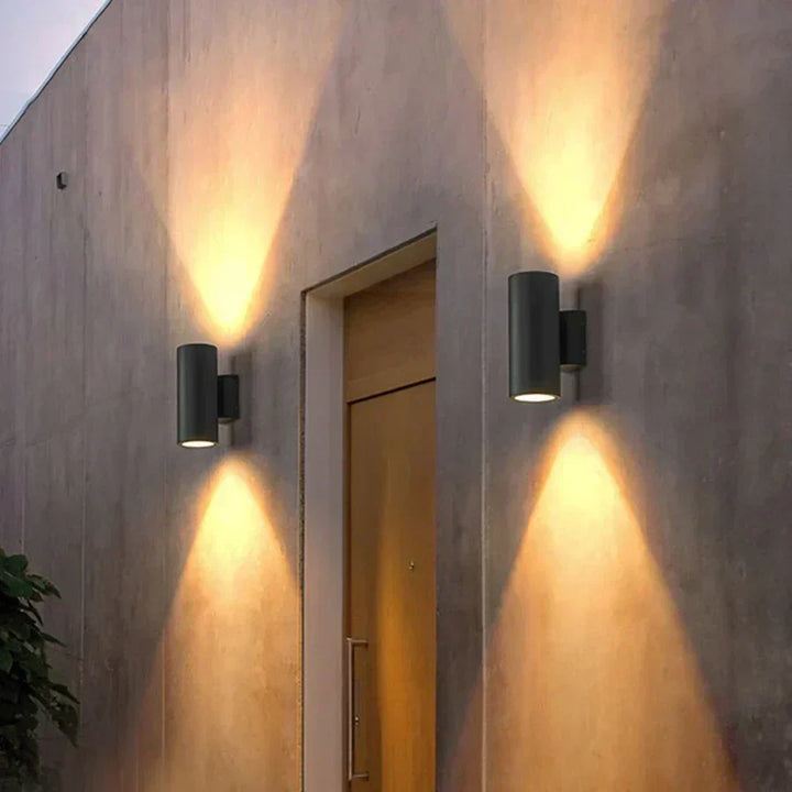Deux appliques cylindriques sombres de part et d'autre d'une porte en bois, dessinant des cônes de lumière chaude sur un mur en béton. 6
