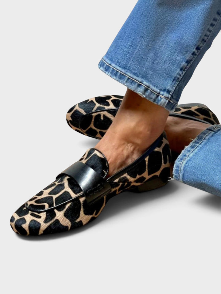 Mocassins à imprimé animalier beige et noir portés avec un jean bleu, pieds croisés, vus de trois quarts avec la bride noire. 1