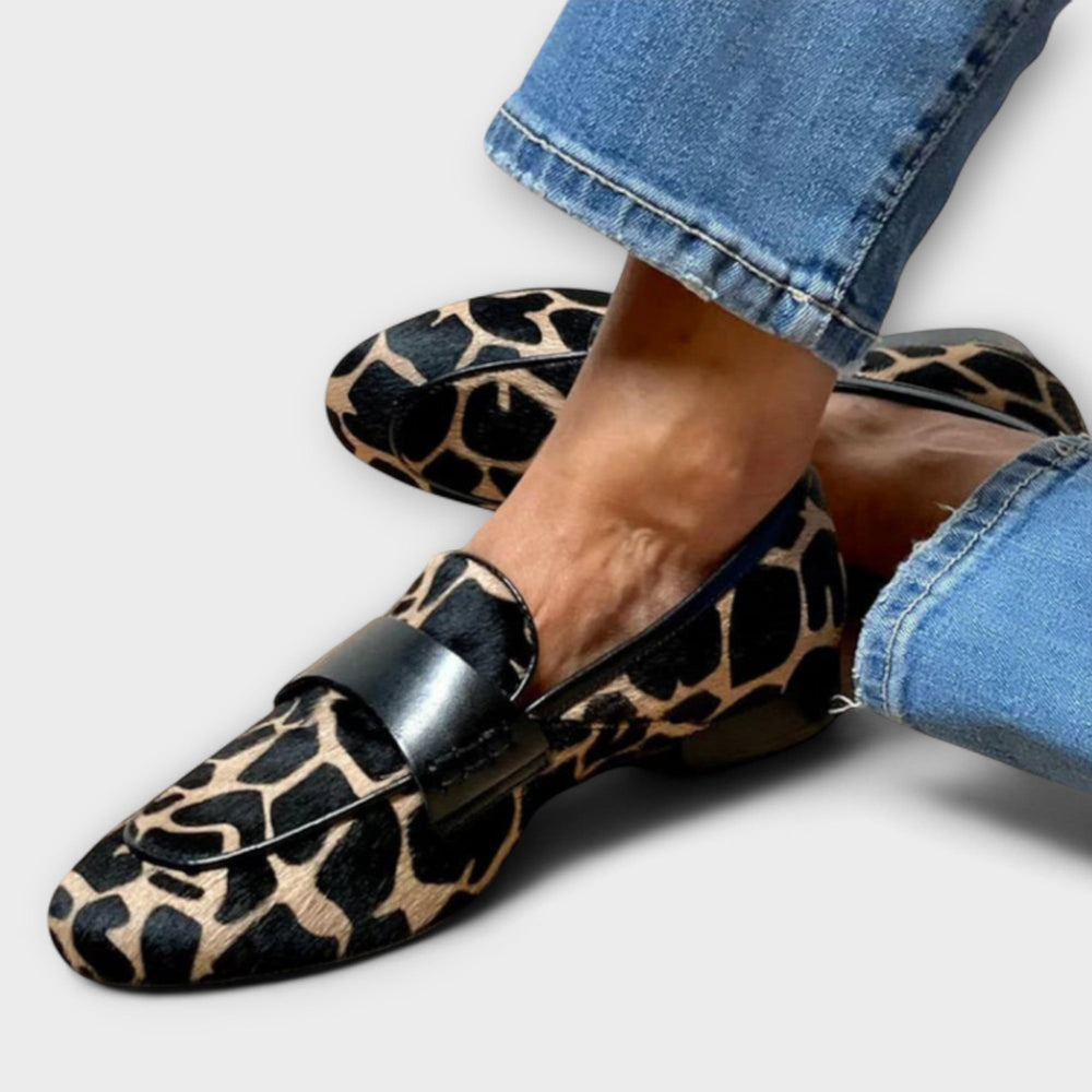 Gros plan de mocassins à imprimé animalier et bride noire en cuir, portés avec un jean bleu retroussé sur fond clair. 2