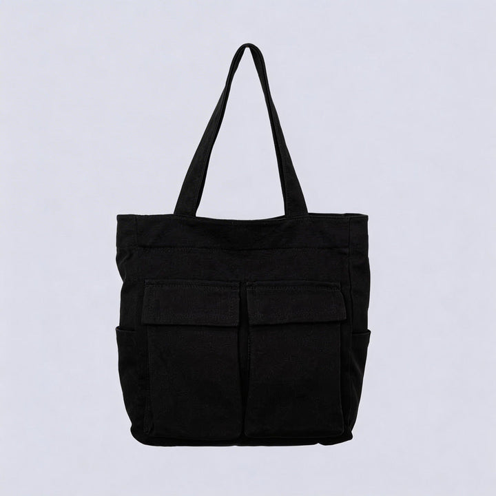 Sac cabas noir en toile, vu de face sur fond clair, avec deux grandes poches à rabat et deux poches latérales. 1