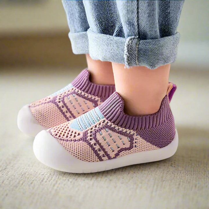 Pieds d'enfant portant des chaussures en maille mauve et rose clair à rayures bleu pâle, vues de côté sur une moquette. 10