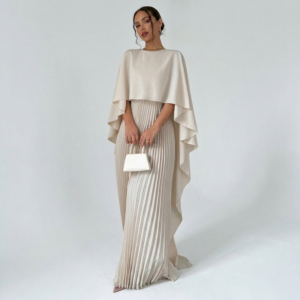 Femme en pied portant la robe beige à cape fluide et jupe plissée, sac blanc à la main 1