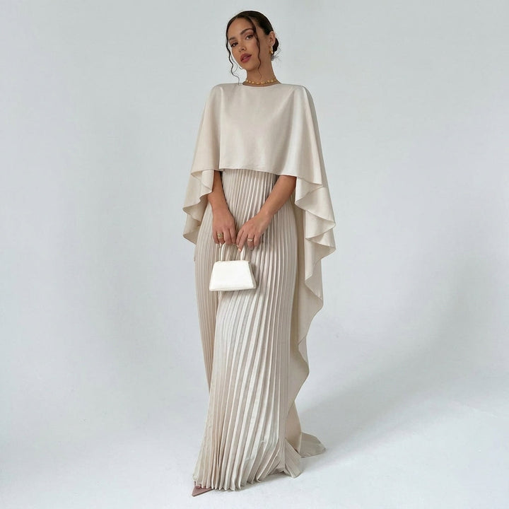 Femme en pied portant la robe beige à cape fluide et jupe plissée, sac blanc à la main 1