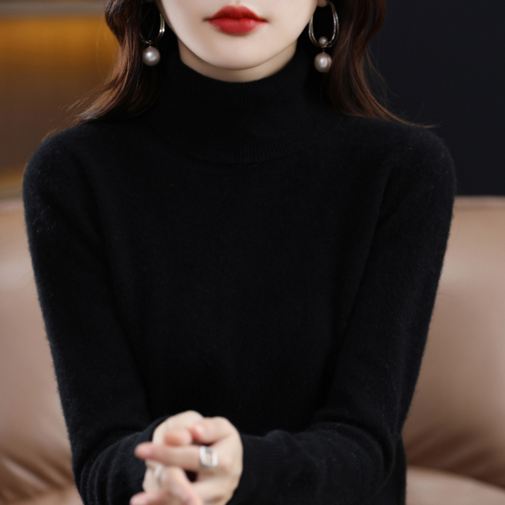 Gros plan sur le buste d'une femme portant le pull noir à col roulé, mains jointes, boucles d'oreilles à perles. 2
