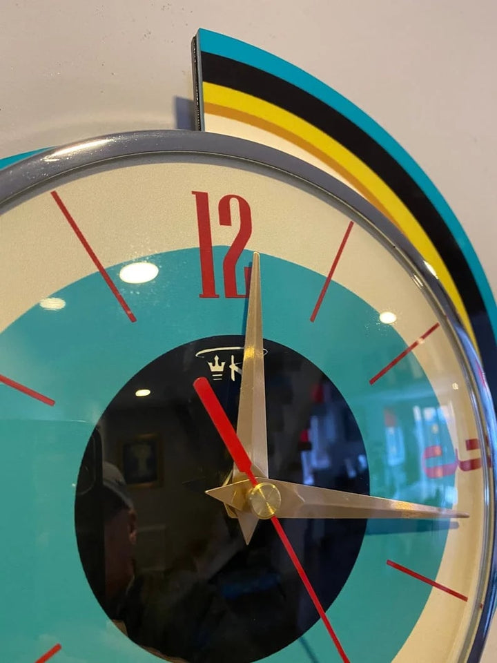 Gros plan sur le cadran de l'horloge spirale: chiffre 12 rouge, aiguilles dorées, trotteuse rouge et verre bombé reflétant la pièce. 10
