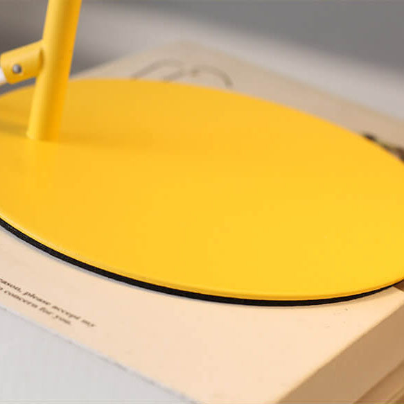 Gros plan sur le socle rond et plat jaune de la lampe, posé sur un livre, avec le pied fin. 4