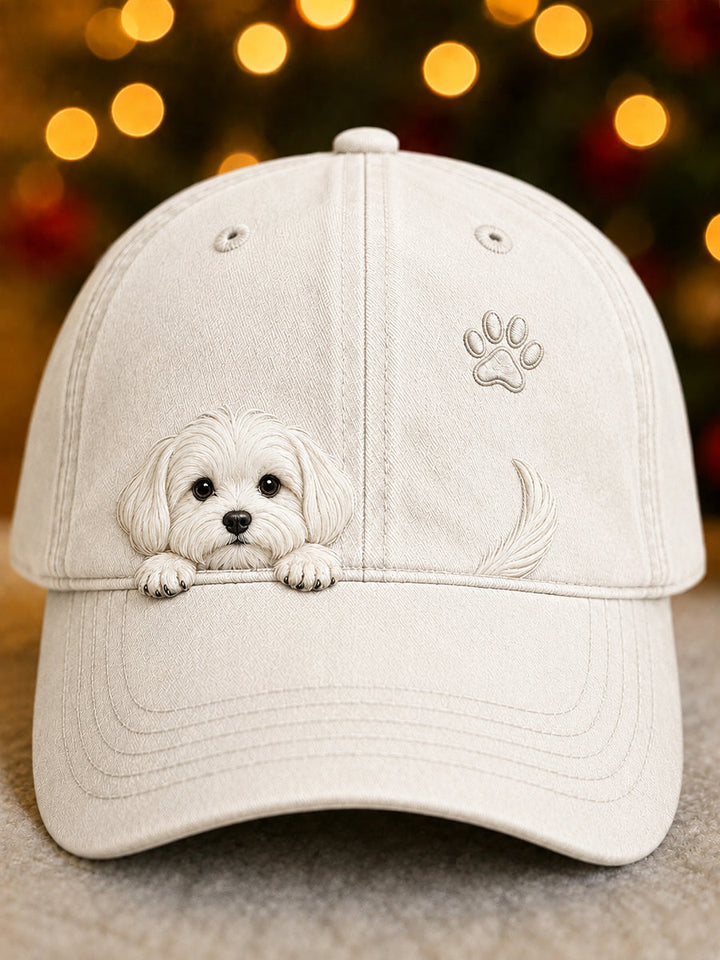 Vue de face de la casquette beige clair ornée d'un chiot blanc en relief, d'une empreinte de patte et d'une queue, devant des lumières de fête. 1
