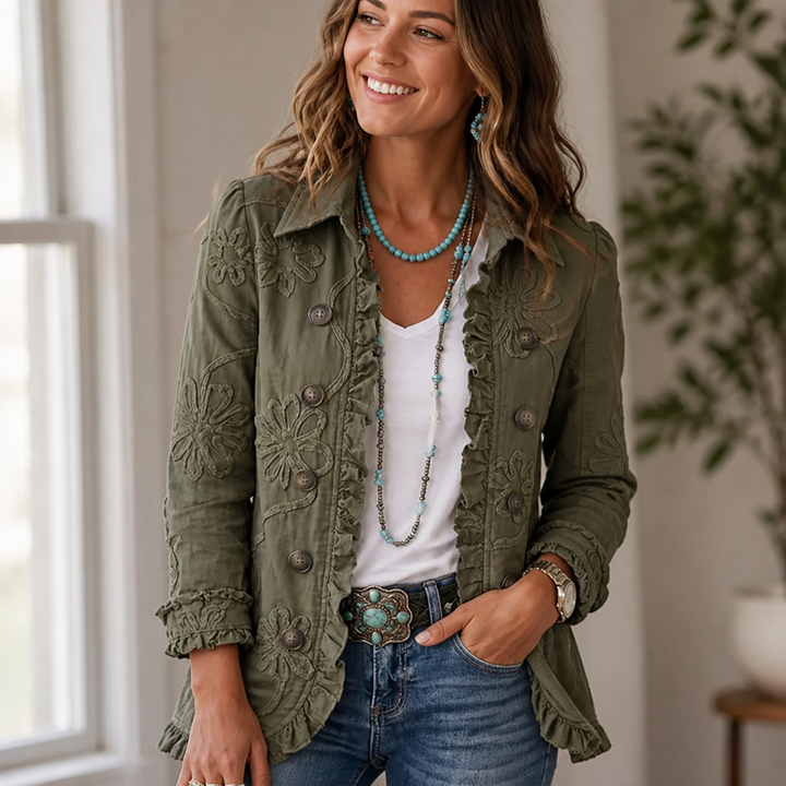 Femme souriante portant la veste kaki brodée de fleurs et bordée de volants, avec t-shirt blanc, jean et colliers turquoise, devant une fenêtre. 1