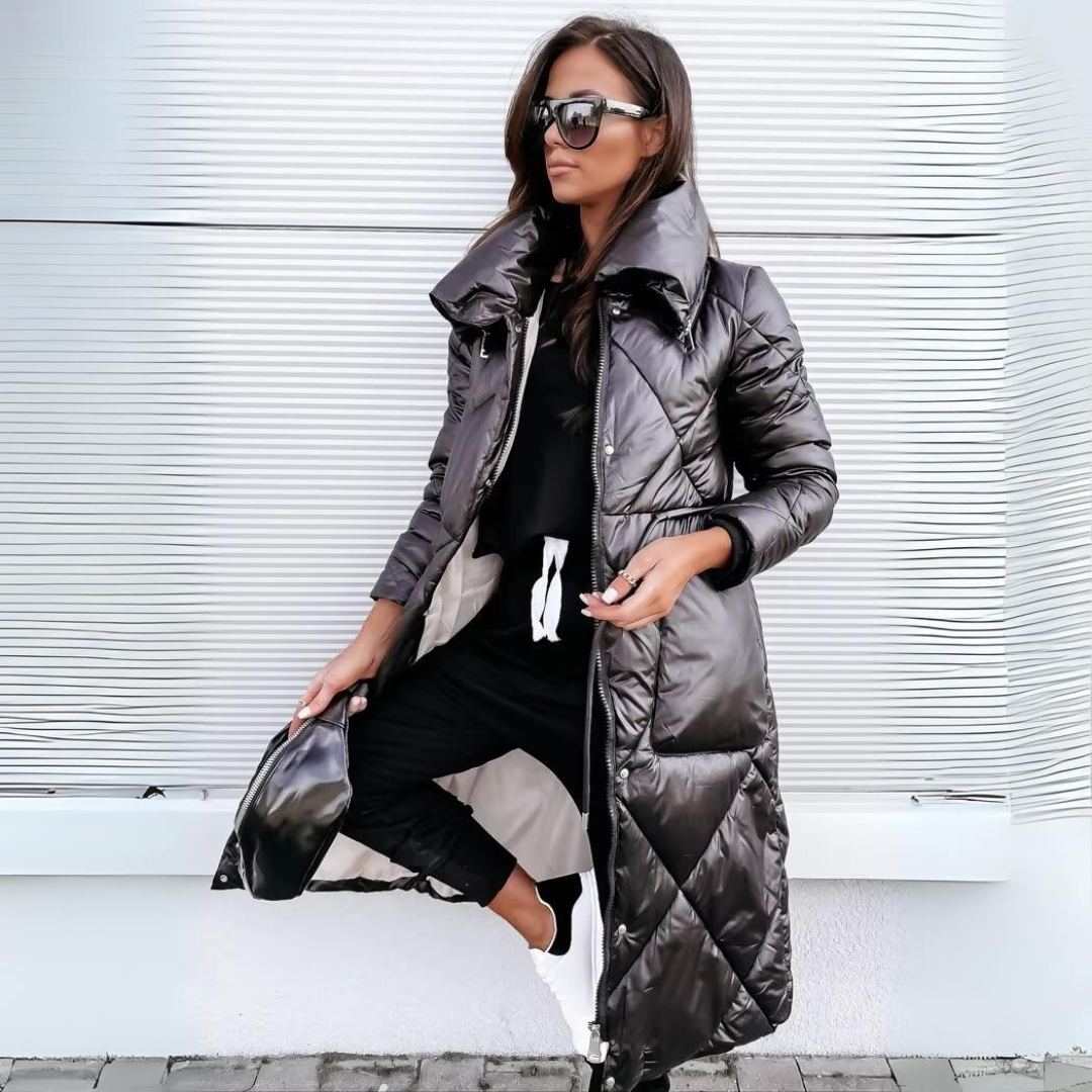 Femme portant le long manteau matelassé noir brillant ouvert, grand col relevé, sur une tenue noire avec baskets blanches, devant un mur blanc. 1