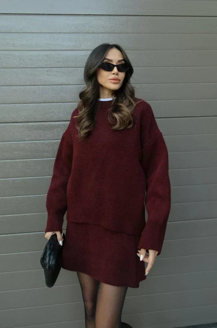 Pull ample et jupe courte évasée en maille bordeaux portés en pied par la mannequin, pochette noire tenue à la main. 2