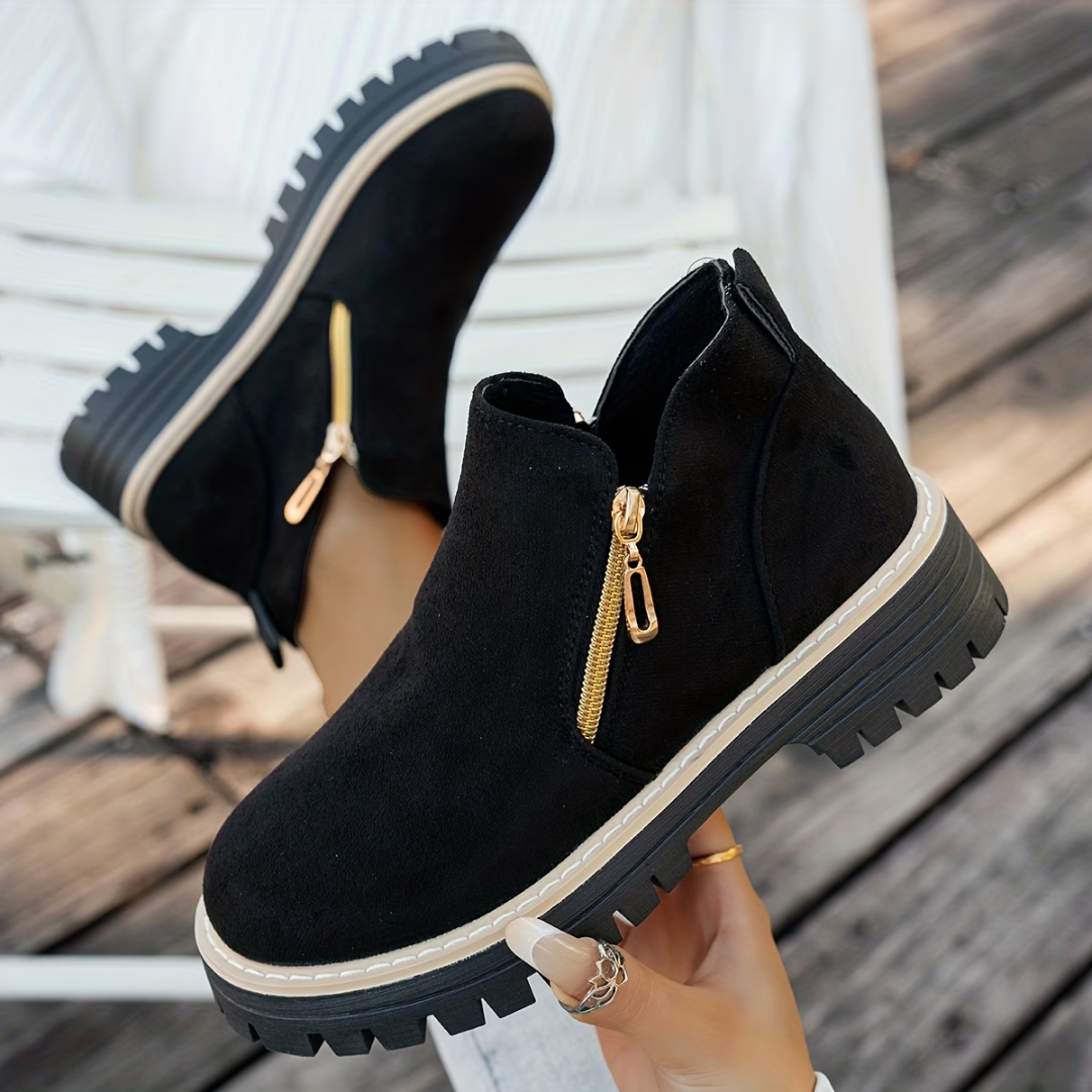 Bottines noires en daim à semelle crantée, fermeture éclair dorée sur le côté, portées pieds croisés sur un banc blanc. 1