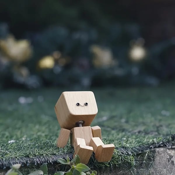 Personnage en bois assis sur une pelouse synthetique, avec un petit sourire, devant un arriere-plan vert flou. 5