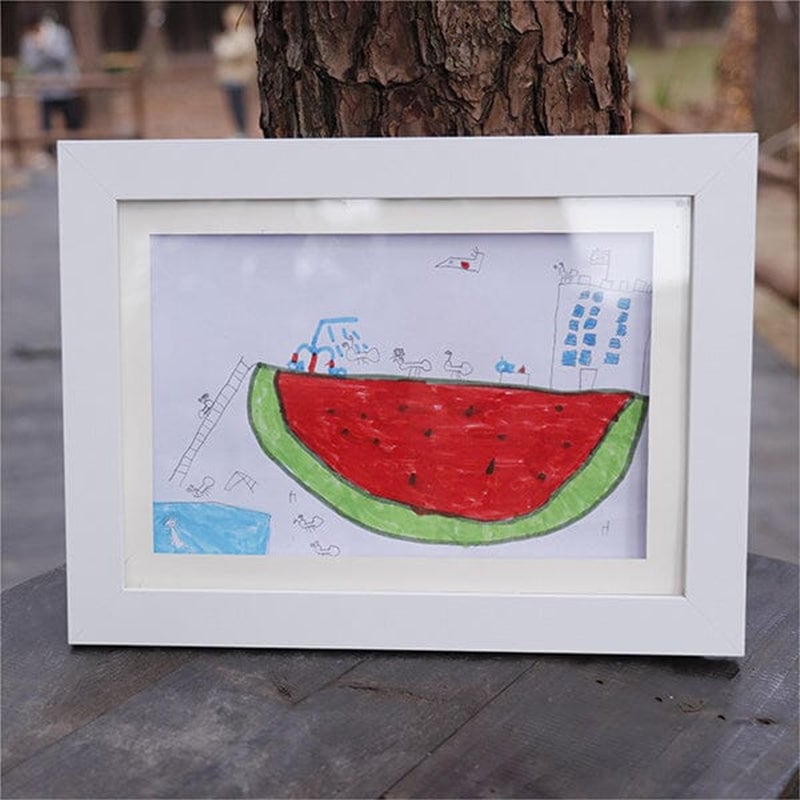 Cadre blanc posé sur une table en bois en extérieur, exposant un dessin d'enfant de pastèque géante au feutre. 21