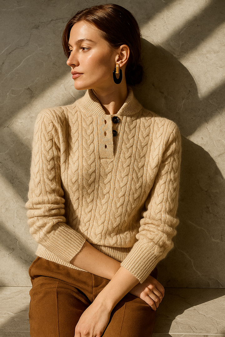 Femme assise contre un mur de pierre, pull beige torsadé à col boutonné, pantalon marron, boucles d'oreilles noires et dorées. 12