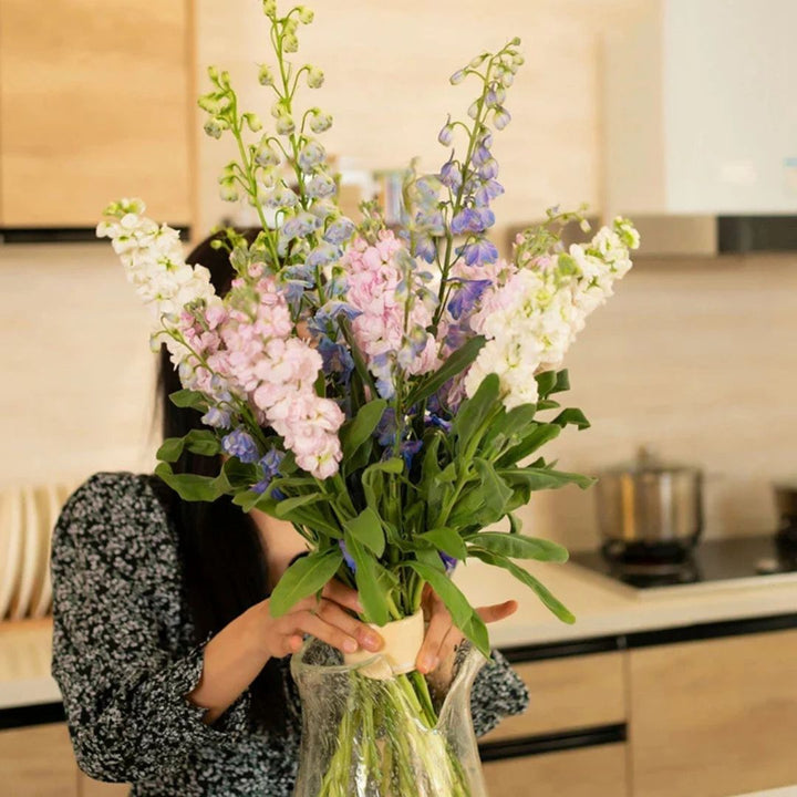 Femme dans une cuisine tenant un haut bouquet de fleurs droites, maintenu par un petit insert beige dans un vase en verre. 2