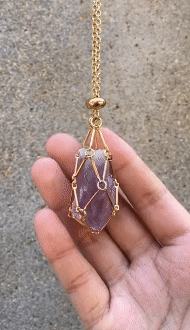Main ouverte portant un collier doré dont le filet retient une pierre violette allongée, sur fond de béton gris. 5