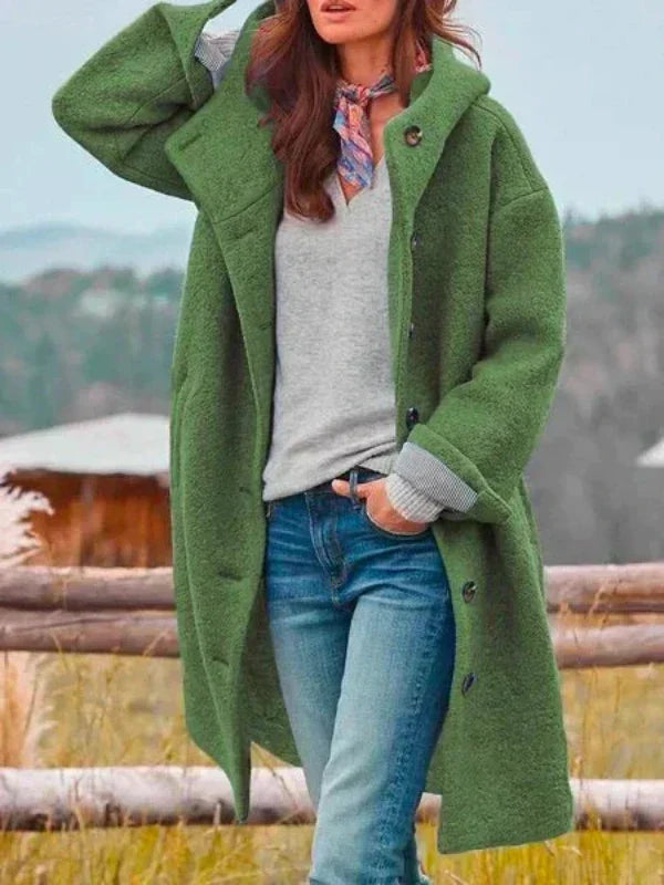 Manteau long à capuche vert porté ouvert avec un pull gris, un jean et un foulard, une main dans la poche. 5