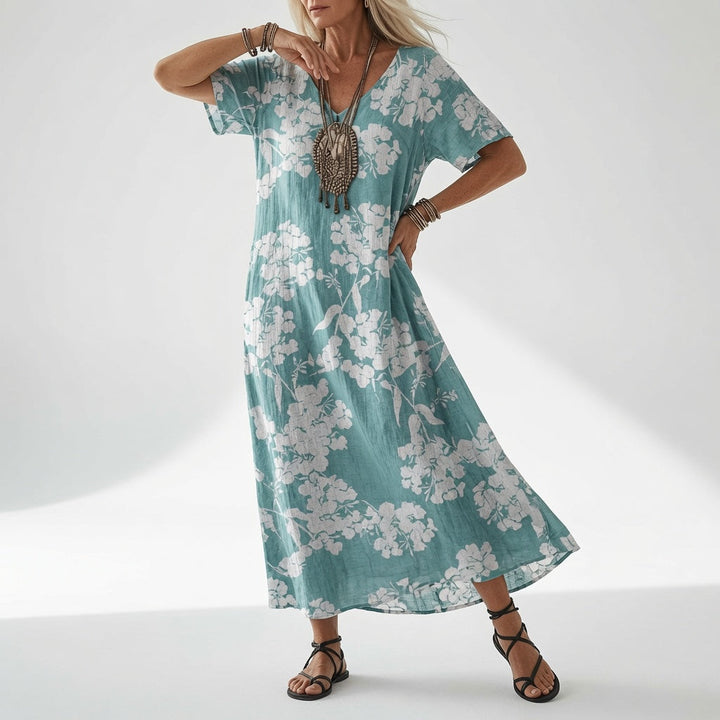 Femme portant la robe fluide bleu turquoise à col en V, imprimée de grappes de fleurs blanches, avec collier et sandales noires. 4