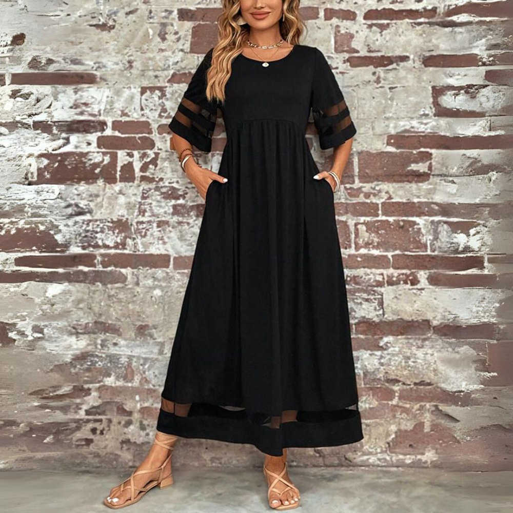 Robe longue noire vue de face, mains dans les poches, devant un mur de briques, avec empiècements transparents aux manches et à l'ourlet. 2