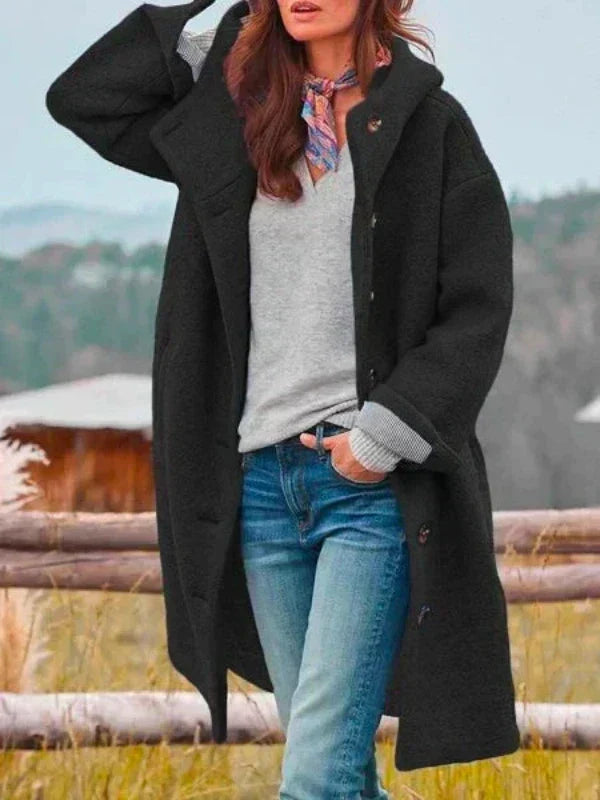Manteau long à capuche noir porté ouvert avec un pull gris, un jean et un foulard coloré, dans un décor champêtre. 3