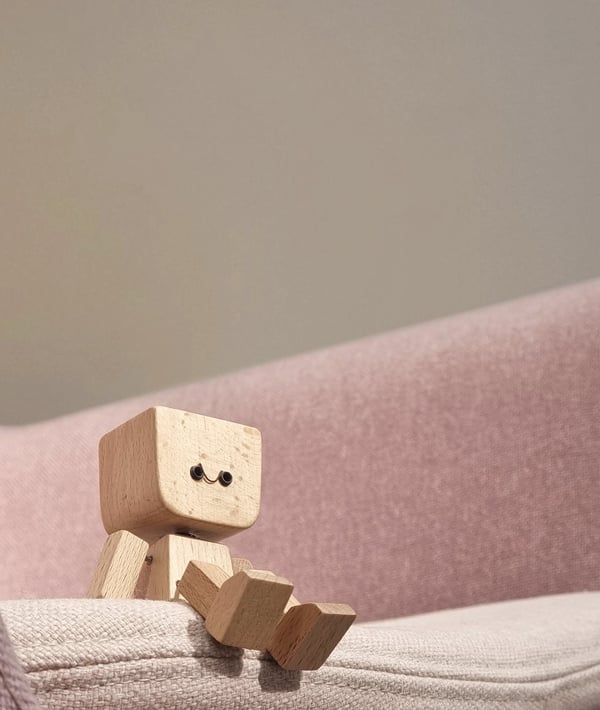 Personnage en bois sourire aux yeux noirs, assis sur le bord d'un fauteuil rose en tissu, jambes tendues. 4