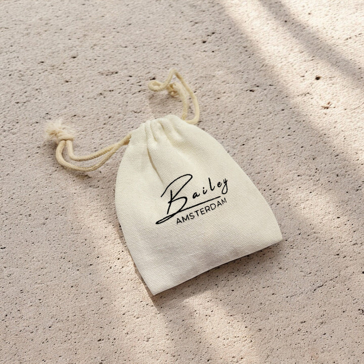 Pochette en tissu beige à cordon posée sur une surface sableuse, emballage du bijou 6