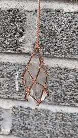Pendentif à filet cuivré retenant une pierre claire, accroché devant un mur de briques grises. 6