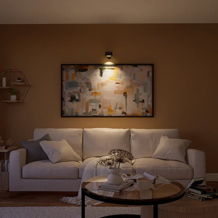 Applique noire éclairant un grand tableau abstrait au-dessus d'un canapé beige, dans un salon aux murs ocre. 8
