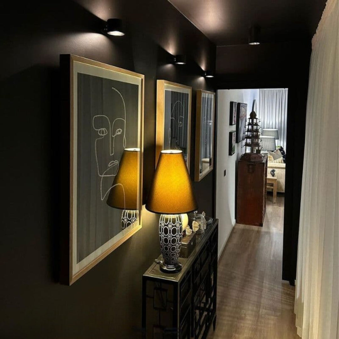 Plusieurs appliques noires fixées dans un couloir sombre, éclairant des cadres au mur et une console avec lampe de table. 10
