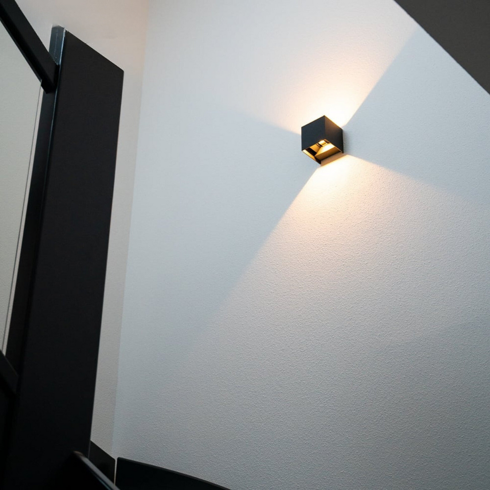 Applique cubique noire vue en contre-plongée dans une cage d'escalier, son éclairage chaud dessinant des cônes de lumière sur un mur blanc. 2
