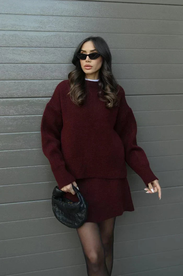 Mannequin portant le pull et la jupe en maille bordeaux, lunettes noires et petit sac tressé noir, debout contre un mur gris. 1