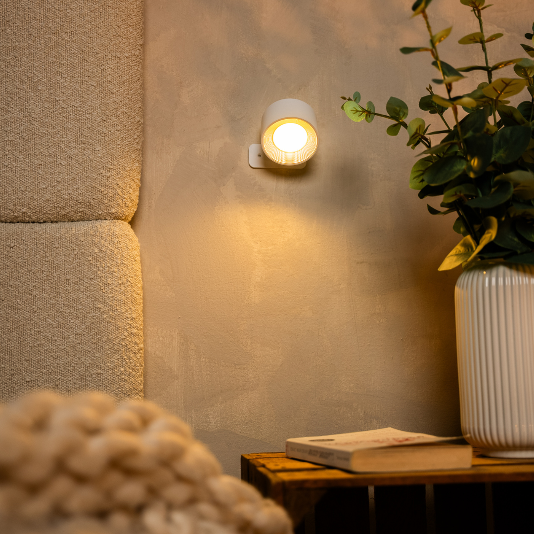 Applique blanche allumée sur un mur enduit, entre un panneau en tissu bouclé et un vase blanc strié avec eucalyptus. 12