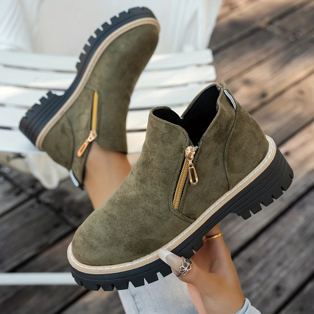 Bottines vert kaki en daim avec zip doré sur le côté et semelle noire crantée, portées sur un banc blanc. 3
