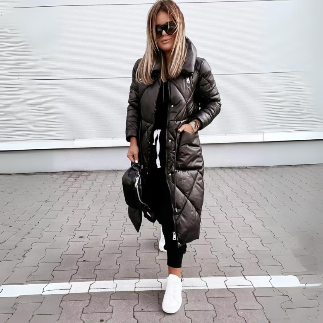Femme blonde en manteau long matelassé noir fermé, une main dans la poche, avec sac noir et baskets blanches sur un pavé. 5