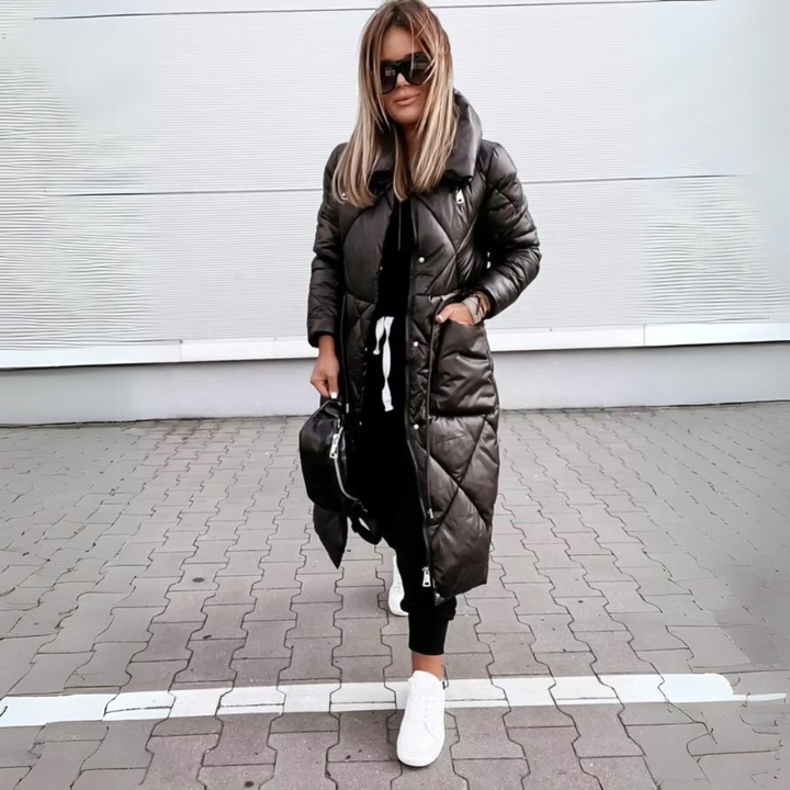 Femme blonde en manteau long matelassé noir fermé, une main dans la poche, avec sac noir et baskets blanches sur un pavé. 5
