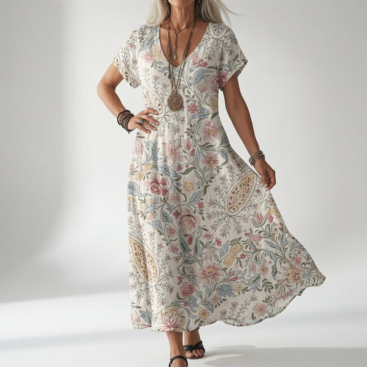 Femme en robe fluide à col en V et motifs floraux et cachemire rose, bleu et jaune sur fond crème, une main sur la hanche. 1