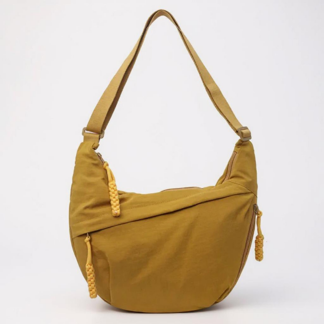 Sac ocre en demi-lune vu de face sur fond clair, avec longue bandoulière réglable et tirettes de zip tressées jaunes. 1